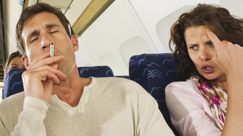 An angry woman looking at a man holding a cigarette on a plane.