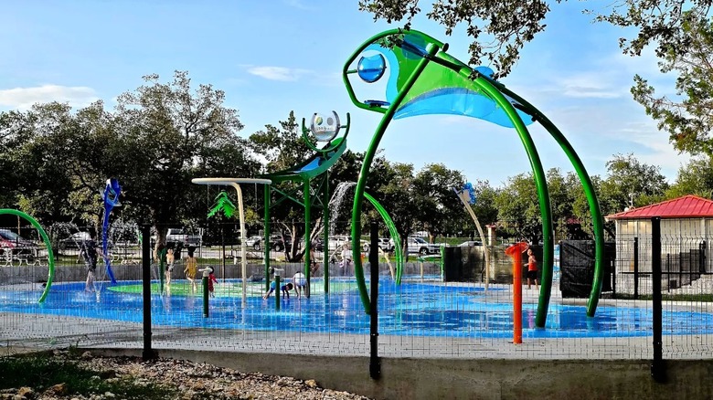 Several kids playing at a splash pad at Brushy Creek Lake Park in Texas, featuring blue and green water jets and spraying fountains.