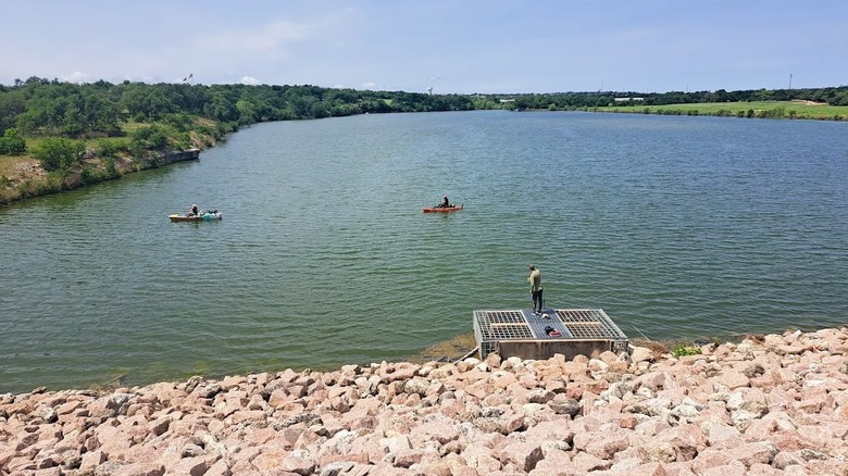 Visitors fishing on a calm lake at Brushy Creek Lake Park near Austin, Texas, featuring a pier,  a rocky embankment, and greenery.