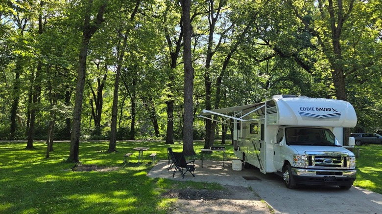 An RV with an extended awning parked beneath a thick canopy of trees at Walnut Woods State Park.