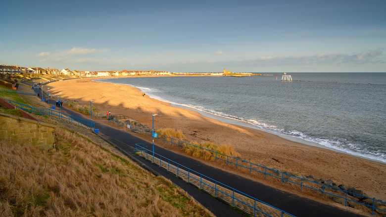 A view of the promenade along Newbiggin Bay, as well as some sandy beach and the famous "The Couple" statue in the distance on a bright day in Newbiggin-by-the-sea, England