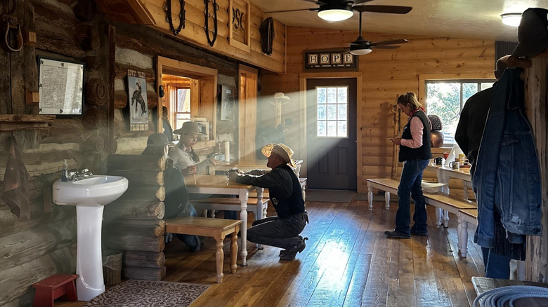 People sit at communal dining tables inside of a log building; light is filtering through the windows