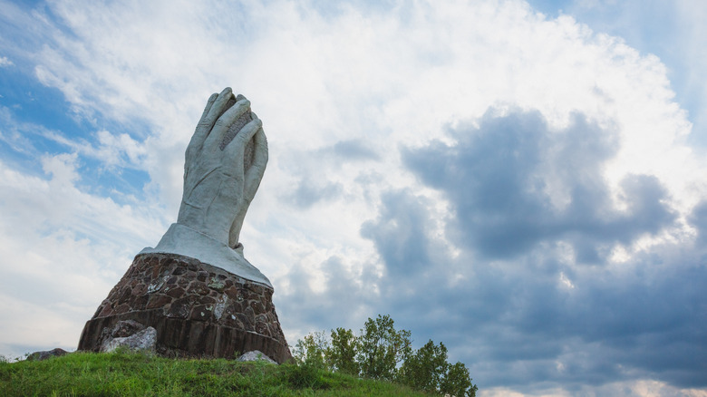 A stucco statue depicting praying hands in King Jack Park in Webb City, set against a cloudy sky