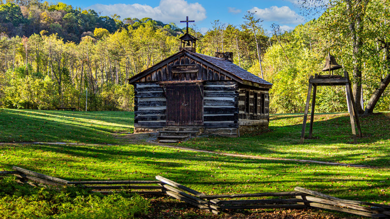 Chapel in the Pioneer Village in Beaver Creek State Park, a one-story log structure with a cross and bell on the roof and a bell on a wooden stand to the right, with a low wood fence in front of them, surrounded by grass with trees in the background under a cloudy blue sky