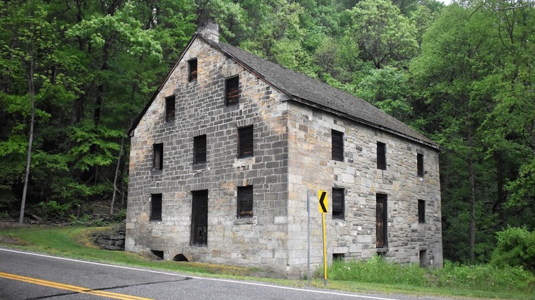 Hambleton's Mill, a stone building with boarded-up windows and doorways next to a road, with a yellow sign beside it and trees in the background
