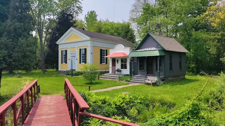 A row of old buildings surrounded by grass and trees with a red bridge in the foreground from the Cobblestone Museum.