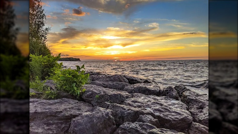 The sun setting over Lake Ontario at Lakeside State Park with rocks in the foreground and trees on the left.
