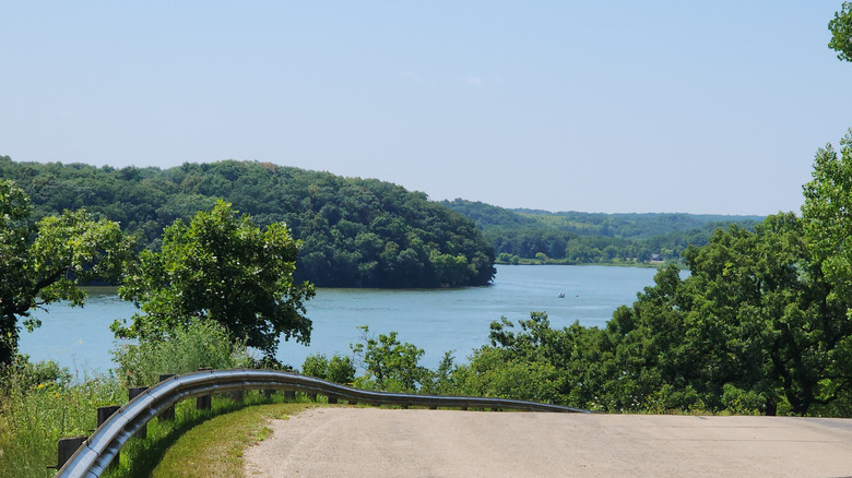 A photo from the road above the lake with a bit of guardrail that turns and the tree-lined lake on the horizon below