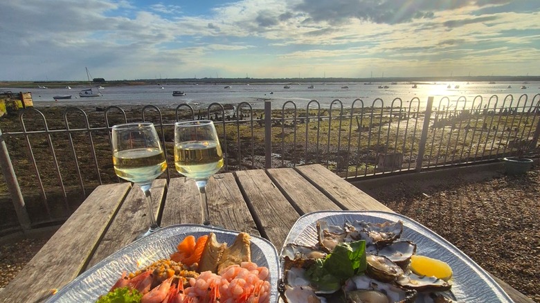 Plates of oysters and prawns and glasses of wine on a picnic table overlooking a boat-filled estuary in West Mersea