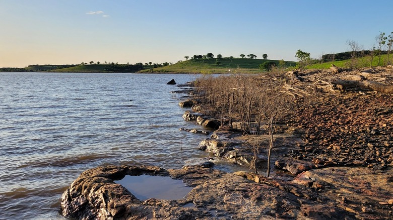Rocky shoreline and distant grassy hills with trees along the calm water at Hulah Lake.