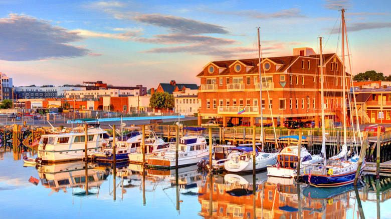 Boats moored at a harbour overlooking brick buildings in Norwalk with the sky reflected in water.