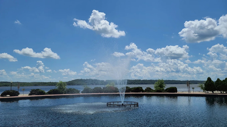 A man-made pond and water fountain sits side-by-side with the Mississippi River in Fort Madison, Iowa under a partly cloudy sky