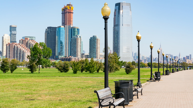 Vast green area lined with street lamps and wooden benches, Jersey City and Manhattan skylines in the distance, Liberty State Park