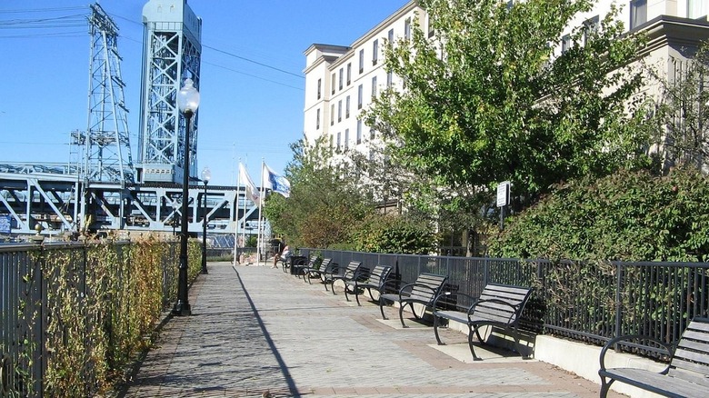 Harrison Riverwalk, a path lined in benches and light posts with a large white building on the right and blue industrial equipment ahead