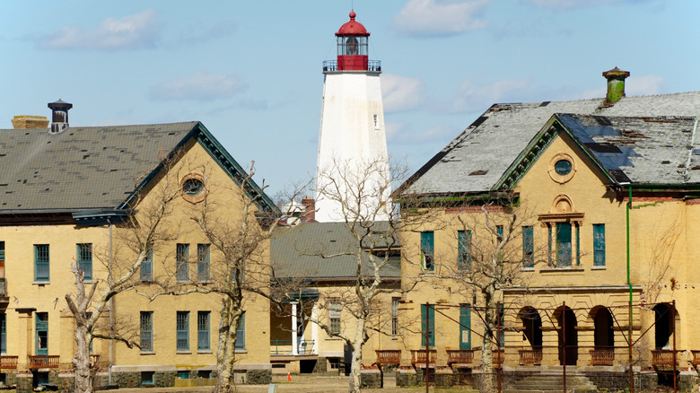Yellow brick buildings with dilapidated gray roofs, with leafless trees in the foreground and the Sandy Hook Lighthouse behind
