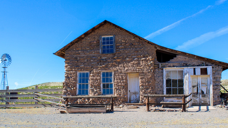 Abandoned stone house next to a small windmill behind a wooden fence, under a blue sky