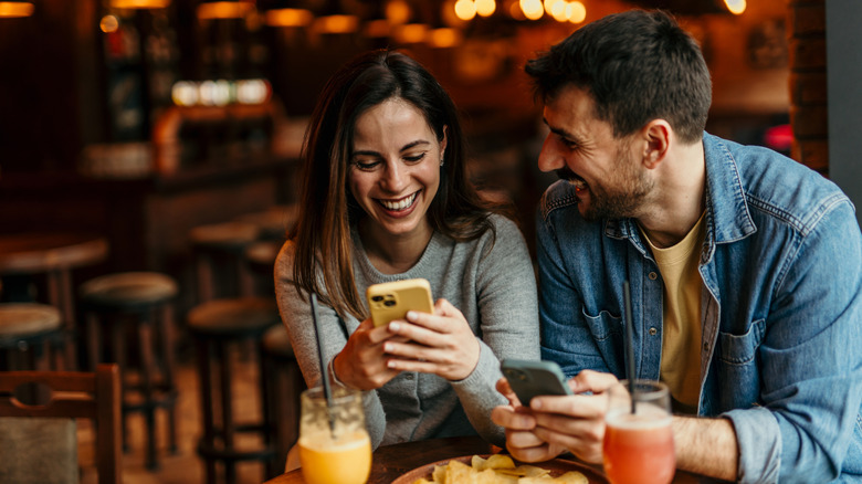 A couple smiling over their phones in front of a table with cocktails and food