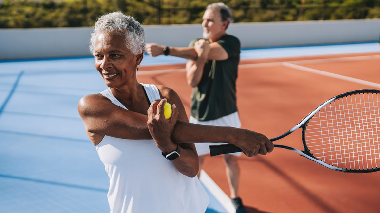 Two senior adults on a tennis court stretching their arms