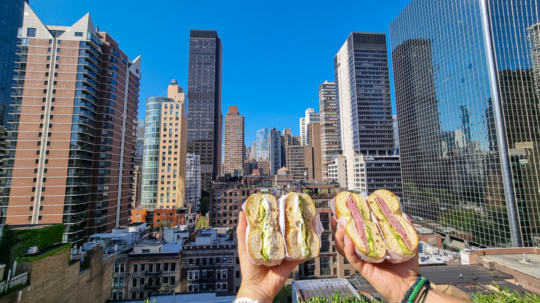Two people hold bagel sandwiches in front of New York skyline