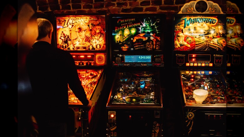 Man plays pinball next to a row of machines in a dark arcade