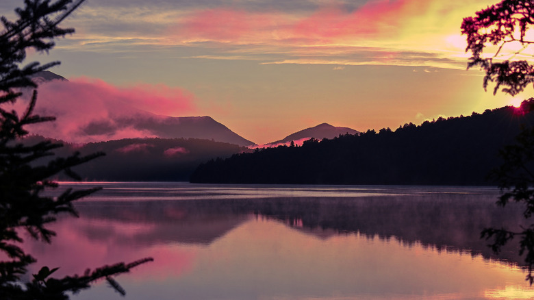 Still lake reflecting sunset colors with backdrop of trees, clouds, and mountains
