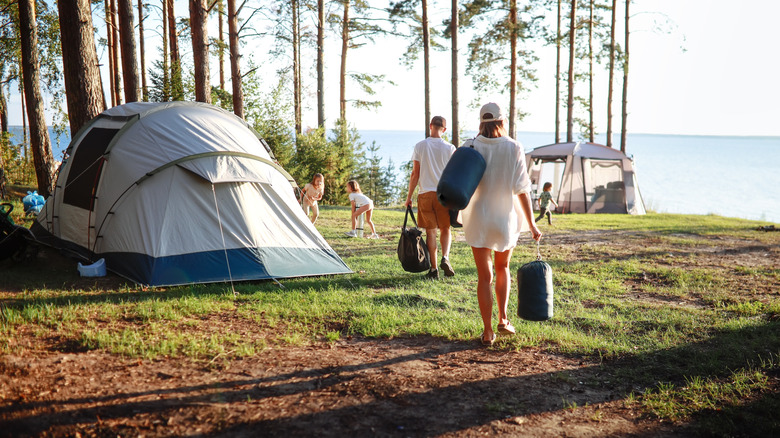 A family dressed in white hold camping equipment as they walk toward their tent and a serene lakeside.