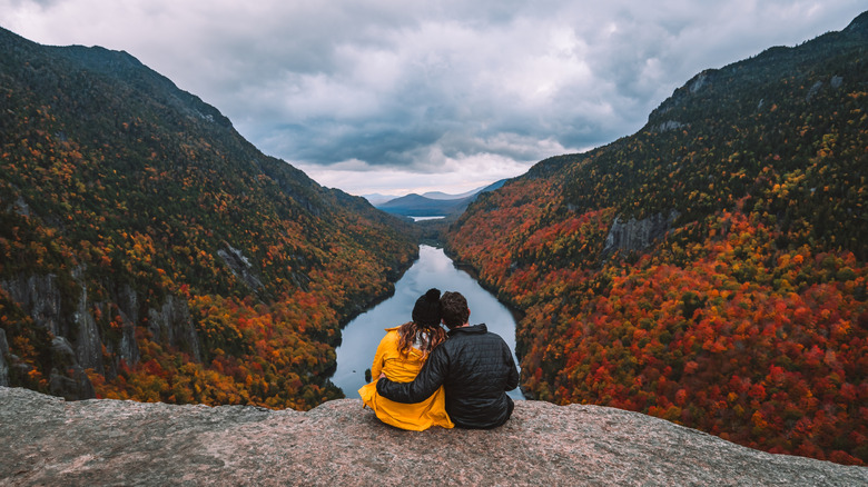 A couple sit on the rocks at Indian Head overlooking a pristine lake and orange-leaf trees along cliffs.