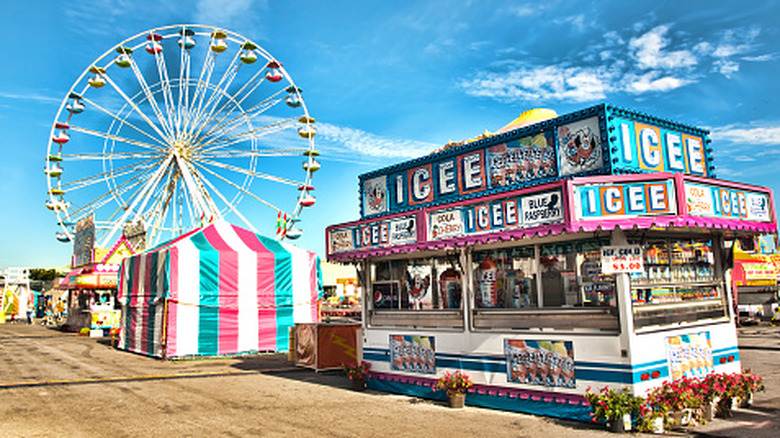 An Icee stand is positioned near a ferris wheel at the Great New York State Fair on a sunny day
