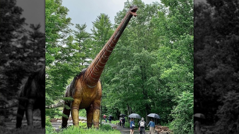 People with umbrellas walking next to an oversize dinosaur figure at Lake George Expedition Park