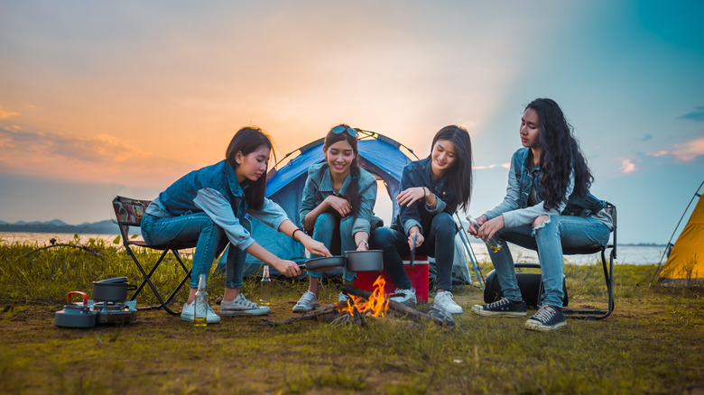 Four campers cook over a fire as the sun sets over a lake behind them