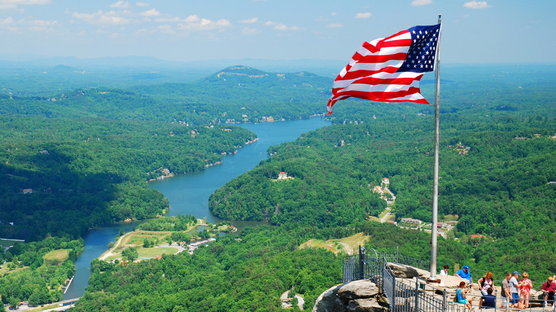 Overlook at Chimney Rock State Park with people on a rocky cliff, a large American flag, and a forested lake below
