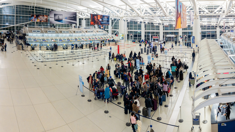 An overhead shot shows a crowd of travelers queuing in front of airline service desks at John F. Kennedy Airport in New York
