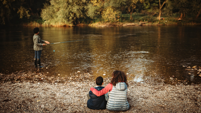 Brother and sister sitting on the side of a lake watching their mother fish.