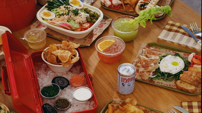 A spread of poolside classic food and drinks from River Electric's Pool Bar is laid out on a wooden table