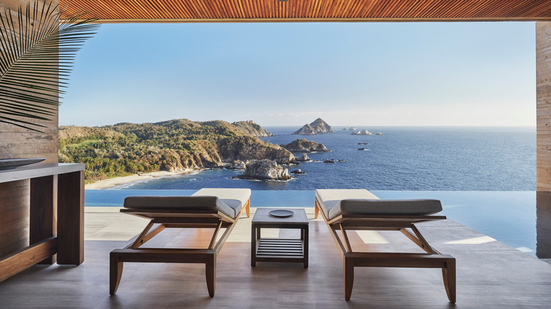 the view is from the covered deck space of the resort's presidential suite, there is an infinity pool lining the outdoor portion of the deck; there are two padded deck loungers in the middle of the image with a small table in between them; in the distance you can see green coastline and small rocky islands;