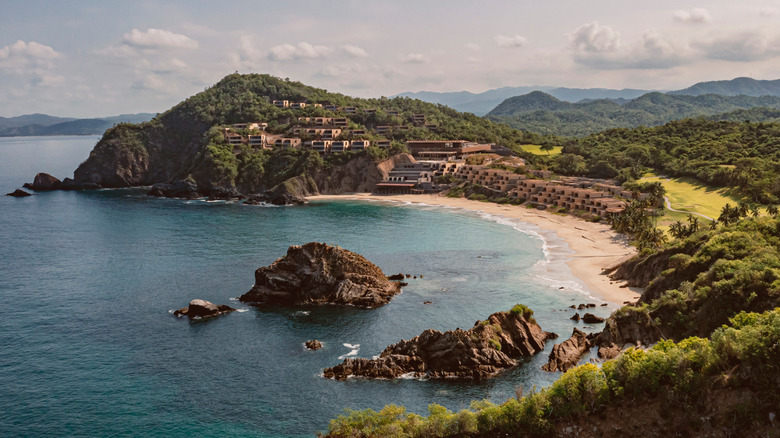 Aerial view of the Four Seasons Resort Tamarindo; the resort sits on a crescent shoreline with the lefthand side of the resort rising up a lush green hill; the resort and its buildings are set into the cliffside and extend down to the beach; behind the resort is a golf course interlaced with tall green trees; in the foreground are a few small rocky islands