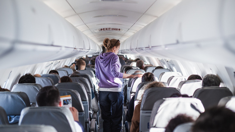 Interior of an airplane from the back as a female traveler wearing a light purple hoodie and navy pants walks in the aisle