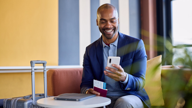 Happy and smiling man using phone while waiting to travel with his suitcase and passport.