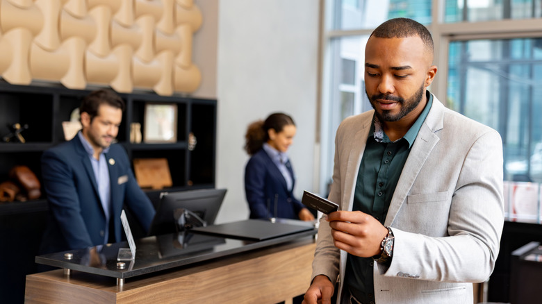 A man in a blazer holding a hotel loyalty card in the lobby of a hotel with staff behind him