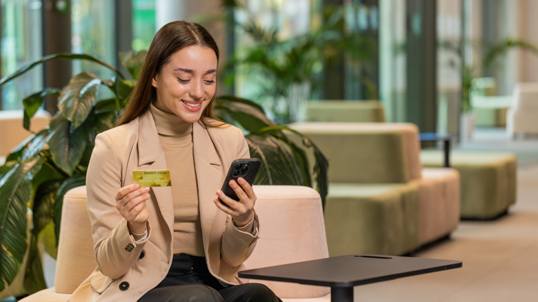 A woman sitting in a hotel lobby, smiling and holding a credit card and phone