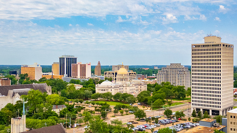 The skyline of Jackson, Mississippi, stands amidst lush green trees and a bright blue sky mottled with white clouds