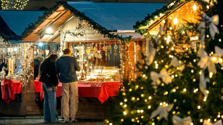 Shoppers stand at a booth selling Christmas ornaments at an outdoor market, with a lighted Christmas tree in the foreground