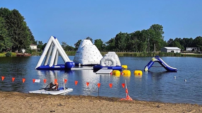 An inflatable obstacle course on the water with a section of beach visible in the foreground and trees and an RV in the background.