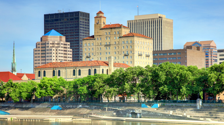 The skyline of Dayton, Ohio, with trees running along cement stairs that lead to the river in the foreground.