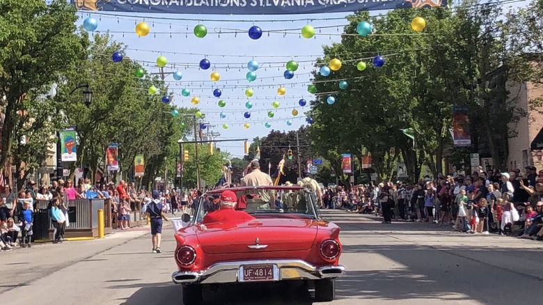 A red car driving down a street lined with tons of people and trees during a community event in Sylvania, OH