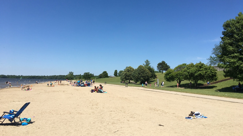 Beach at Buck Creek State Park, with a blue lawn chair in the foreground and people on the sand, trees and grass along the right of the frame, and the lake along the left of the frame, under a blue sky