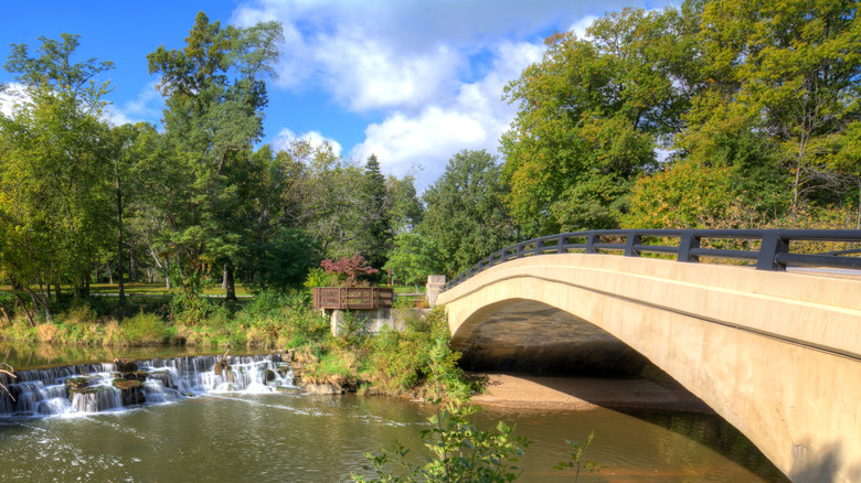 Baldwin Lake in Mill Stream Run Reservation, with a tan bridge with a black railing on the right, a small waterfall on the left, and green trees in the background under a cloudy blue sky