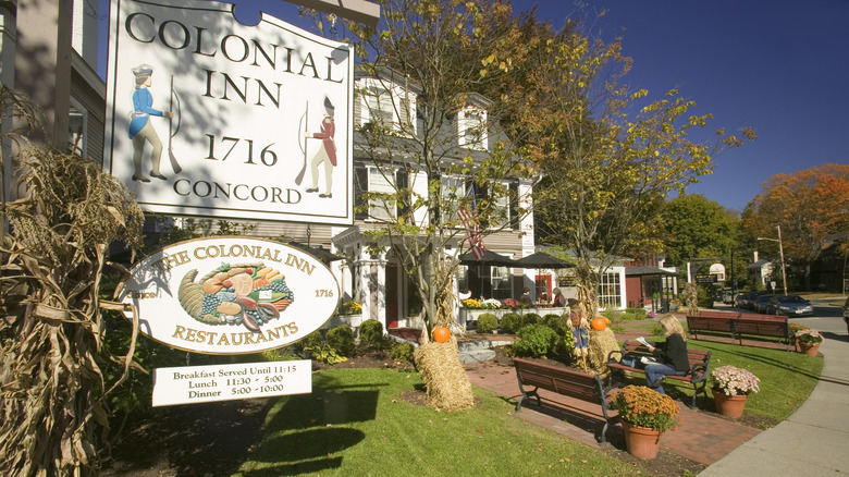 Sign of hte Colonial Inn, with the gray-roofed white front of the inn in the background and benches lining a brick walk through grass, under a blue sky