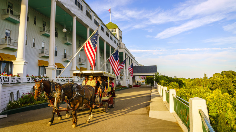 Horse-drawn carriage in front of Grand Hotel, a white building with tall columns, with green trees in the right of the frame under a cloudy blue sky