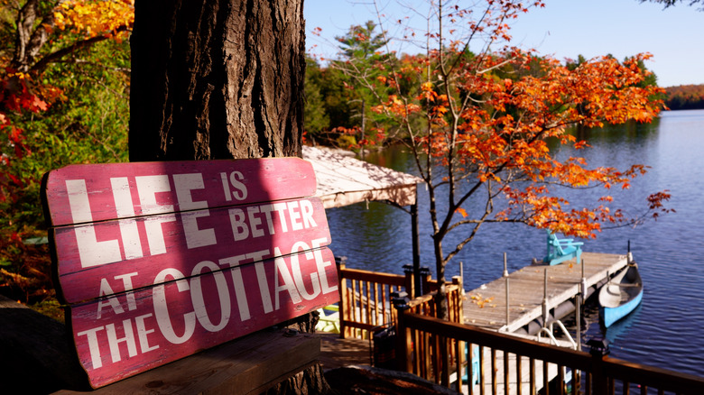 A sign reading "Life is better at the cottage" among tress and a lake setting